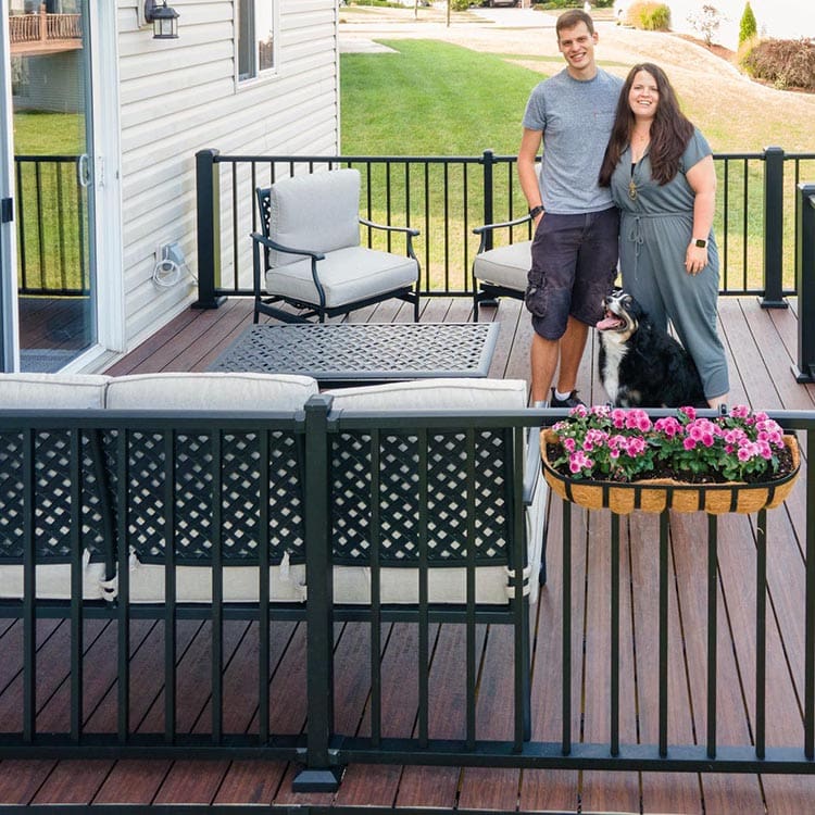 A couple standing on their new deck that's adorned with outdoor sofas and a chair and flowers