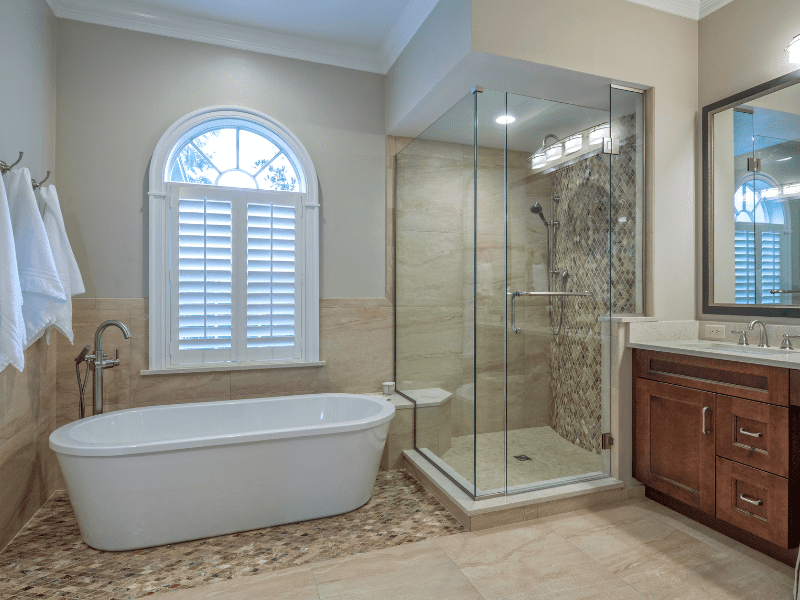 An earth-tone bathroom with travertine floors, porcelain tile squares under the stand-alone tub and in the glassed-in. walk-in shower; wooden vanity and wood framed mirror. A large window with a plantation shutter above the tub.