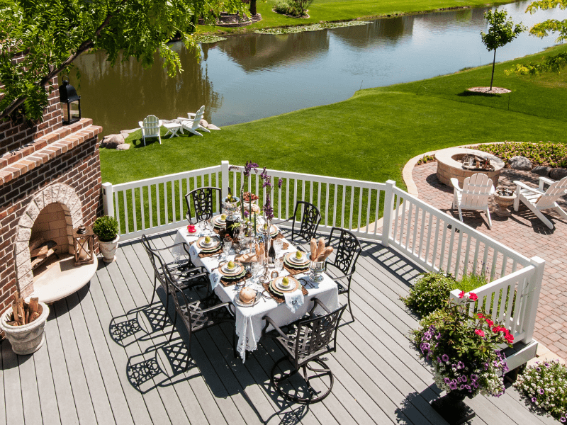 A large deck with a fireplace, a set dining table for 6, overlooking a canal and a green, grassy yard.
