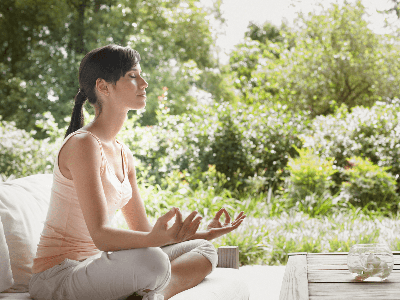 A young woman with a ponytail sitting in lotus position on her deck doing yoga. Lots of greenery and plant life behind her.