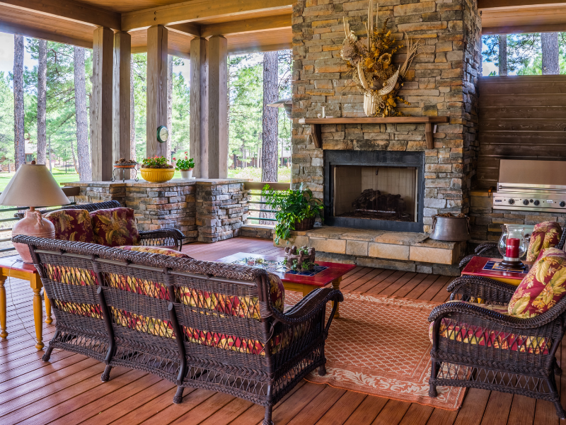 A large redwood deck with a stone fireplace, stone edging, and screened-in sides. A comfy 3-person sofa and two chairs of wicker and cushions face the fireplace.