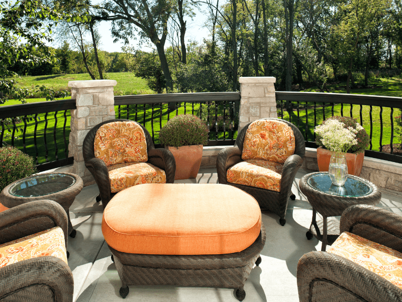 A close-up of an octagonal deck overlooking a green wooded area with metal railings, stone pillars, and wicker chairs and an ottoman. Cushions are organe and floral.