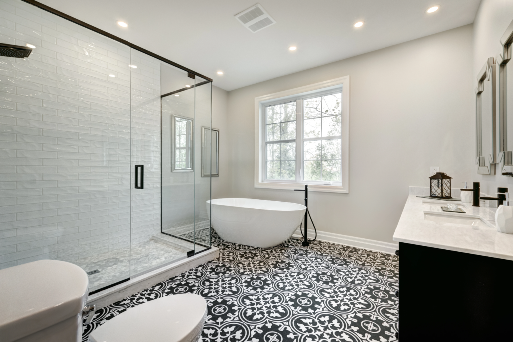 A large bathroom with deco-patterned black, grey, white mosaic tile, a walk-in glass shower, a black vanity with dual sinks, and a stand-alone soaker tub.

