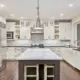A view of a modern, white, sleek kitchen from one end, island in the center, Carera marble countertops, pendent lights over the island.